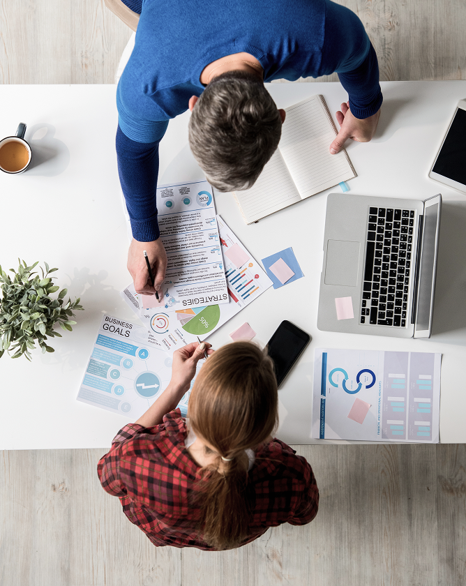 Overhead view of two professionals reviewing strategy documents, business goals, and charts on a desk with a laptop and coffee.